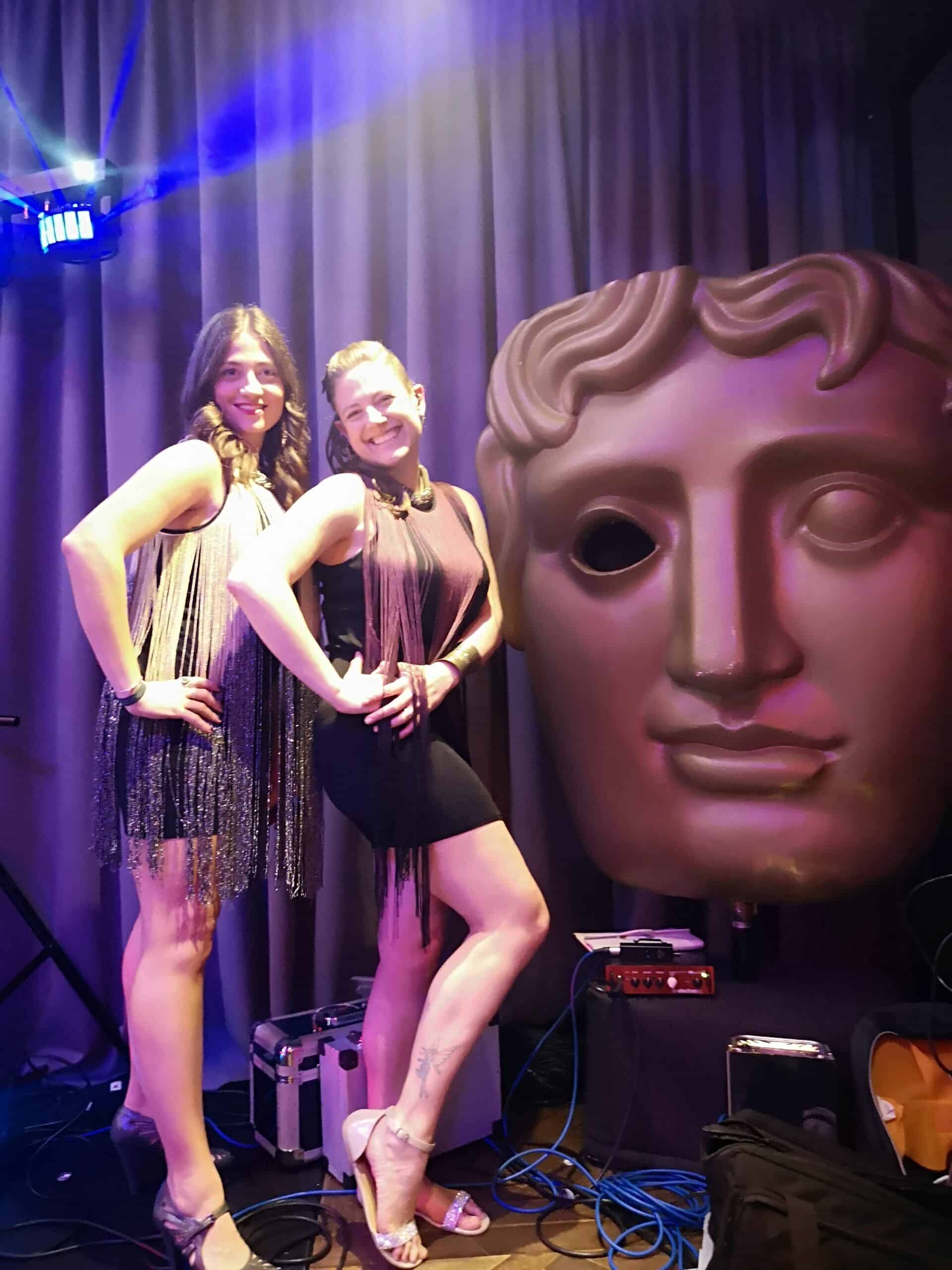 Two women posing beside large BAFTA mask at event
